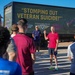 A group of people are gathered outdoors in front of a large truck bearing a 'Stomping Out Veteran Suicide!' billboard from the Veteran Trike Brigade organization. A man in a red shirt appears to be addressing the group with a raised fist, while another man in a blue shirt stands nearby. The setting appears to be a parking area with mountains visible in the background.