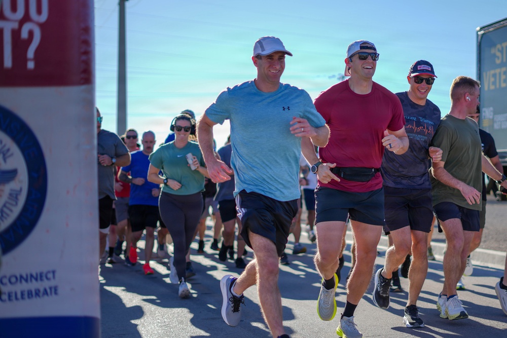 A group of runners participating in what appears to be an organized road race or charity run event. The foreground features three men running together, smiling and appearing to enjoy the event. Partial signage visible on the left references words like 'CONNECT,' 'CELEBRATE,' and 'VETERAN,' suggesting a veterans-themed or military-affiliated event.