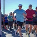 A group of runners participating in what appears to be an organized road race or charity run event. The foreground features three men running together, smiling and appearing to enjoy the event. Partial signage visible on the left references words like 'CONNECT,' 'CELEBRATE,' and 'VETERAN,' suggesting a veterans-themed or military-affiliated event.