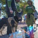 A black and tan German Shepherd wearing a tactical/service harness stands at an outdoor public event, appearing alert and happy with mouth open. Several people dressed in matching green shirts are gathered in the background, suggesting an organized group event or walk. Shopping bags from Natural Grocers and other brands are visible on the ground near the dog.