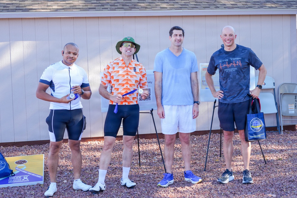 Four men are posing together outdoors near a building, appearing to be at a triathlon or athletic event. Two of the men are holding medals or awards, and one is carrying a branded tote bag. The setting includes gravel ground, folding chairs, and a visible 'Suicide Prevention' awareness sign in the lower left corner.