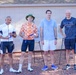 Four men are posing together outdoors near a building, appearing to be at a triathlon or athletic event. Two of the men are holding medals or awards, and one is carrying a branded tote bag. The setting includes gravel ground, folding chairs, and a visible 'Suicide Prevention' awareness sign in the lower left corner.