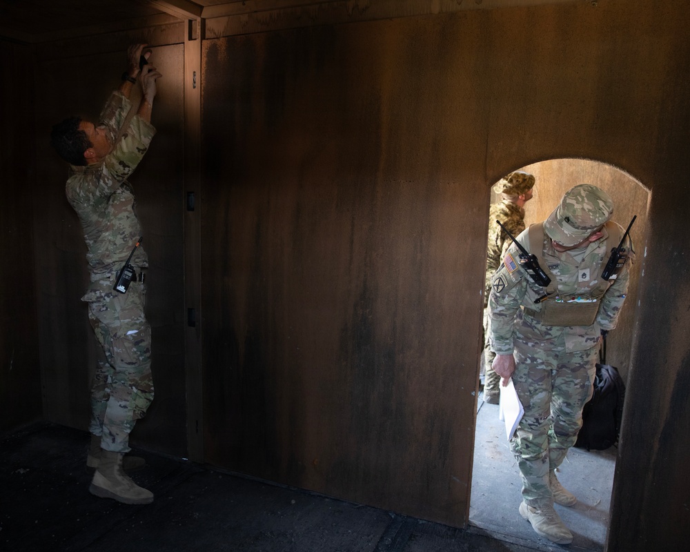 Two or three U.S. Army soldiers in MultiCam/OCP camouflage uniforms are conducting what appears to be a training exercise inside a mock building or shoot house. One soldier on the left is reaching up toward the top of a wooden wall or door, while another soldier enters through an arched doorway holding papers and carrying two radios. The setting appears to be a military training facility with plywood-constructed rooms.