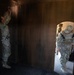 Two or three U.S. Army soldiers in MultiCam/OCP camouflage uniforms are conducting what appears to be a training exercise inside a mock building or shoot house. One soldier on the left is reaching up toward the top of a wooden wall or door, while another soldier enters through an arched doorway holding papers and carrying two radios. The setting appears to be a military training facility with plywood-constructed rooms.