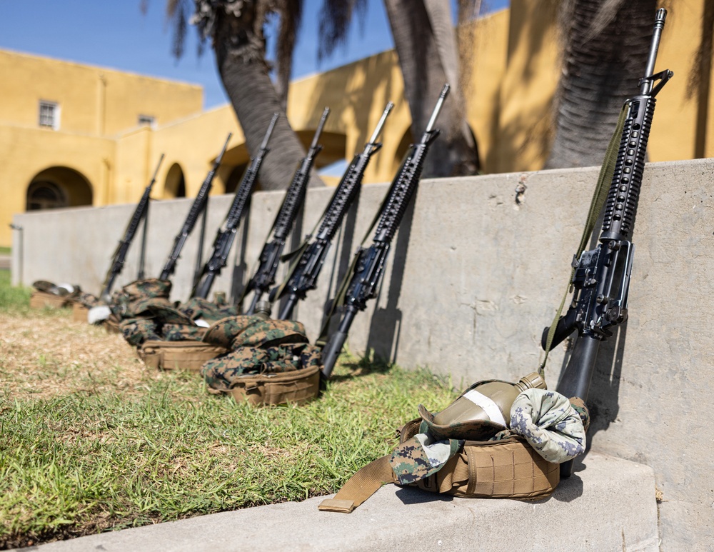 A row of military rifles leaning against a concrete wall outdoors, accompanied by tactical gear and camouflage equipment on the ground. The setting appears to be a military base or installation, identifiable by the yellow stucco buildings and palm trees in the background. The gear includes MARPAT (Marine pattern) camouflage items consistent with U.S. Marine Corps equipment.