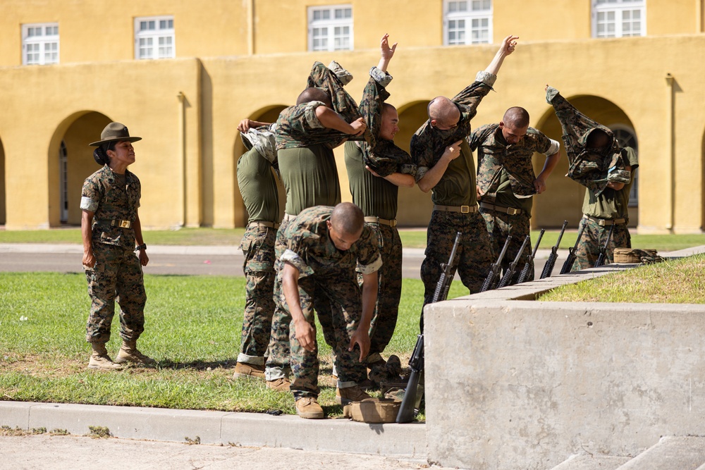 A group of military personnel in USMC woodland camouflage uniforms are engaged in what appears to be a physical training or inspection activity on a parade ground. Several recruits are raising their arms and having their shirts lifted, suggesting a uniform or body inspection, while a drill instructor in a campaign cover (drill sergeant hat) observes from the left. The setting appears to be a Marine Corps base, identifiable by the characteristic yellow stucco building with arched colonnades in the background.