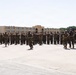 A large formation of military personnel in camouflage uniforms is assembled on a parade deck or drill field. Several individuals appear to be running or moving along the front of the formation, likely drill instructors or cadre. The setting appears to be a military base with a multi-story institutional building and palm trees visible in the background, consistent with a Southern California installation.