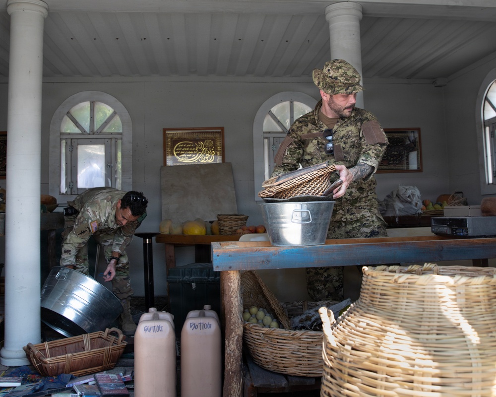 Two military personnel in camouflage uniforms are searching or inspecting what appears to be a market stall or shop space. One soldier wearing a US flag patch bends down examining items on the floor, while another in a different camouflage pattern handles wicker baskets on a table. The setting includes Arabic calligraphy on the wall, suggesting a location in a Middle Eastern or Central Asian country.