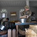 Two military personnel in camouflage uniforms are searching or inspecting what appears to be a market stall or shop space. One soldier wearing a US flag patch bends down examining items on the floor, while another in a different camouflage pattern handles wicker baskets on a table. The setting includes Arabic calligraphy on the wall, suggesting a location in a Middle Eastern or Central Asian country.