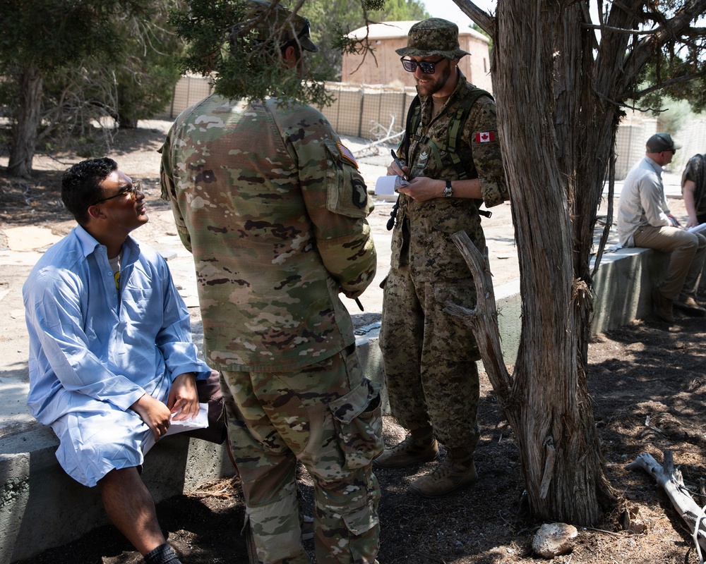 The image shows a military interaction scene where two soldiers — one in US Army OCP (Operational Camouflage Pattern) uniform and one in Canadian Armed Forces CADPAT uniform with a visible Canadian flag patch — are speaking with a civilian man seated on a concrete barrier. The setting appears to be an outdoor area with trees, dirt ground, and what looks like a forward operating base or training environment in the background. Additional personnel are visible in the background engaged in similar activities.