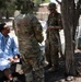 The image shows a military interaction scene where two soldiers — one in US Army OCP (Operational Camouflage Pattern) uniform and one in Canadian Armed Forces CADPAT uniform with a visible Canadian flag patch — are speaking with a civilian man seated on a concrete barrier. The setting appears to be an outdoor area with trees, dirt ground, and what looks like a forward operating base or training environment in the background. Additional personnel are visible in the background engaged in similar activities.