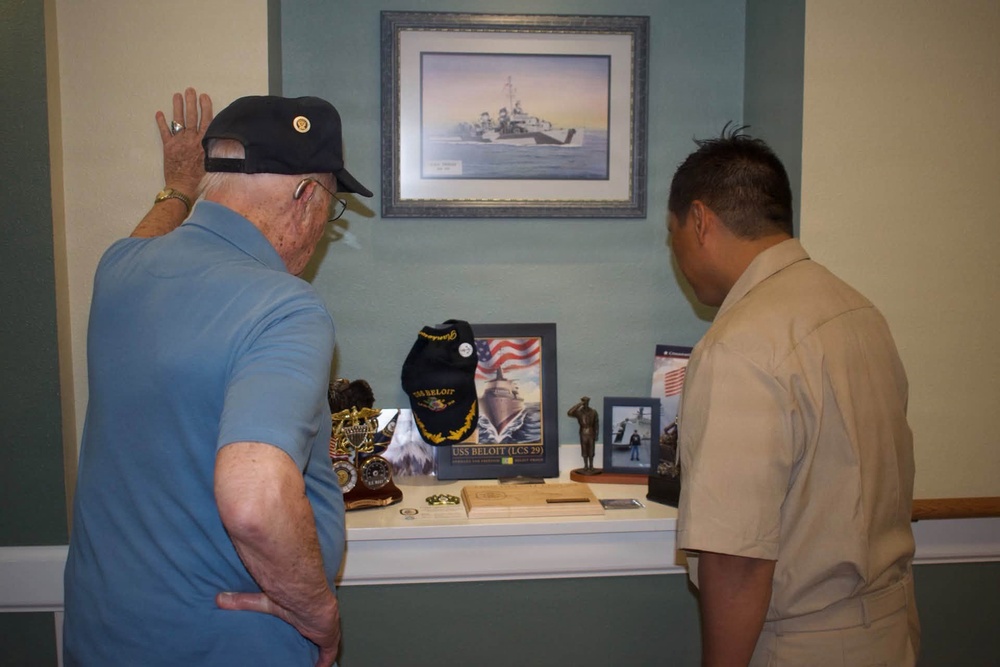 Two individuals are examining a military memorial display shelf featuring USS Beloit (LCS 29) memorabilia. An elderly man wearing a Navy veteran cap raises his hand toward a framed painting of a naval vessel mounted on the wall above the display. The display contains various naval artifacts, photographs, and commemorative items.