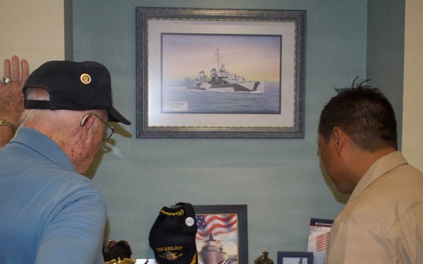 Two individuals are examining a military memorial display shelf featuring USS Beloit (LCS 29) memorabilia. An elderly man wearing a Navy veteran cap raises his hand toward a framed painting of a naval vessel mounted on the wall above the display. The display contains various naval artifacts, photographs, and commemorative items.