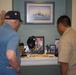 Two individuals are examining a military memorial display shelf featuring USS Beloit (LCS 29) memorabilia. An elderly man wearing a Navy veteran cap raises his hand toward a framed painting of a naval vessel mounted on the wall above the display. The display contains various naval artifacts, photographs, and commemorative items.