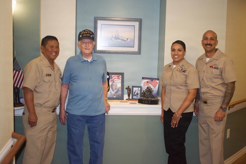 Four individuals pose together in what appears to be a care facility or veterans' home hallway. Three are wearing U.S. Navy khaki uniforms, while one elderly man in civilian clothes wears a veteran's cap. Behind them is a display shelf with military memorabilia and a framed naval ship artwork on the wall.