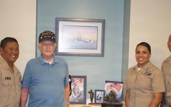 Four individuals pose together in what appears to be a care facility or veterans' home hallway. Three are wearing U.S. Navy khaki uniforms, while one elderly man in civilian clothes wears a veteran's cap. Behind them is a display shelf with military memorabilia and a framed naval ship artwork on the wall.