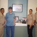 Four individuals pose together in what appears to be a care facility or veterans' home hallway. Three are wearing U.S. Navy khaki uniforms, while one elderly man in civilian clothes wears a veteran's cap. Behind them is a display shelf with military memorabilia and a framed naval ship artwork on the wall.