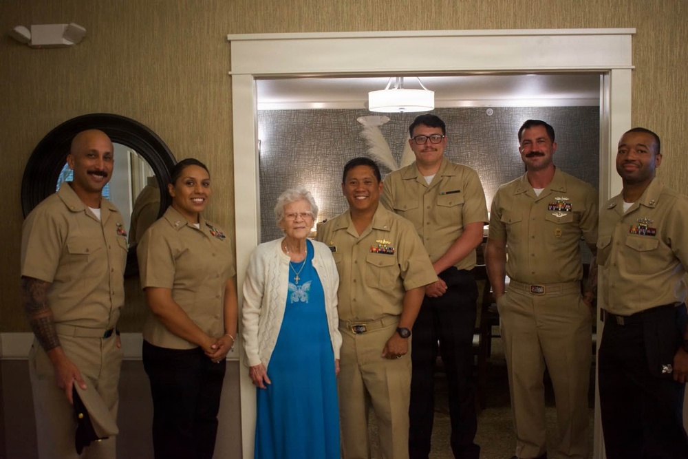 A group of six U.S. Navy personnel in tan/khaki service uniforms are posing for a photo with an elderly civilian woman dressed in a blue dress and white cardigan. The setting appears to be an indoor facility, possibly a senior living or care center, suggesting this may be a community outreach or visitation event. The group is smiling and arranged in a line for a formal group portrait.