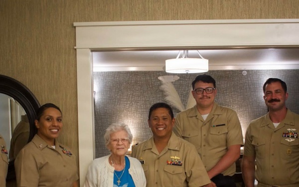 A group of six U.S. Navy personnel in tan/khaki service uniforms are posing for a photo with an elderly civilian woman dressed in a blue dress and white cardigan. The setting appears to be an indoor facility, possibly a senior living or care center, suggesting this may be a community outreach or visitation event. The group is smiling and arranged in a line for a formal group portrait.