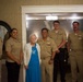 A group of six U.S. Navy personnel in tan/khaki service uniforms are posing for a photo with an elderly civilian woman dressed in a blue dress and white cardigan. The setting appears to be an indoor facility, possibly a senior living or care center, suggesting this may be a community outreach or visitation event. The group is smiling and arranged in a line for a formal group portrait.