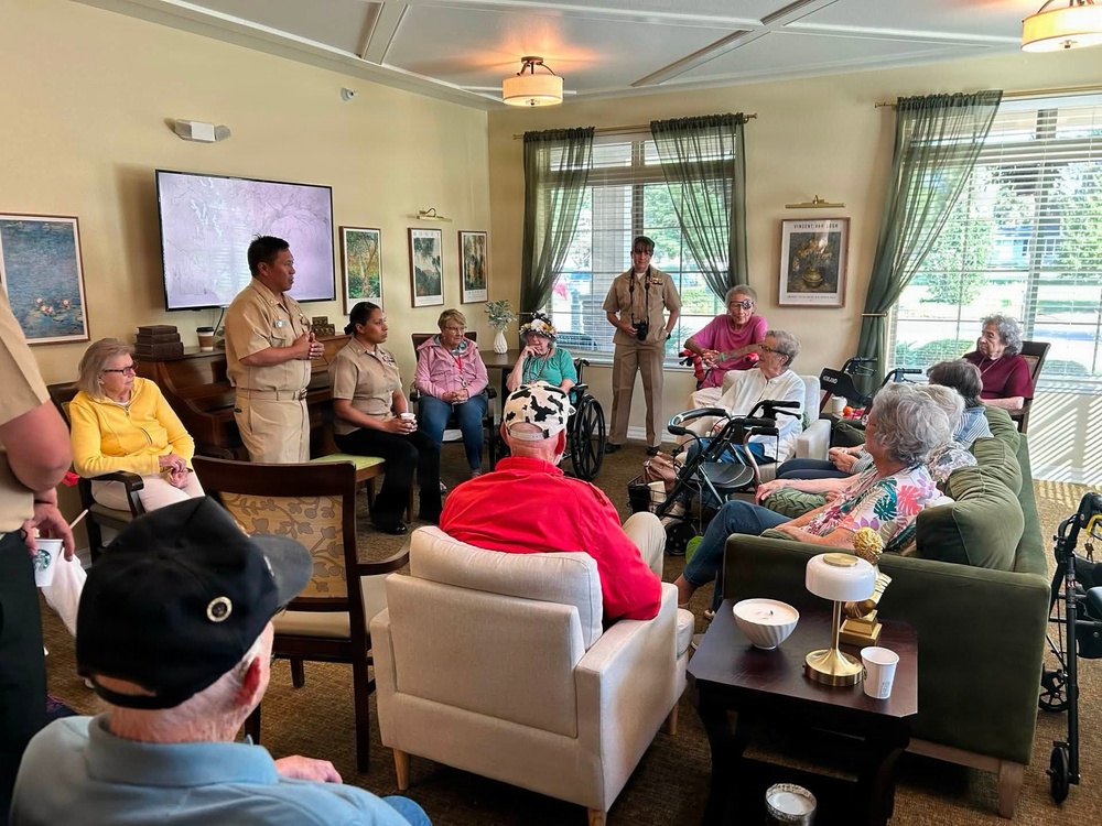 Military personnel in tan/khaki uniforms are conducting a presentation or visit at what appears to be a senior living facility or assisted living community. A service member stands near a display screen while addressing a group of elderly residents seated in a comfortable lounge-style room. The setting suggests a community outreach or veterans appreciation event.
