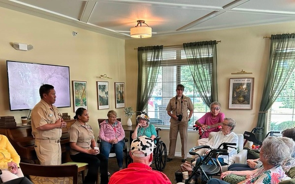Military personnel in tan/khaki uniforms are conducting a presentation or visit at what appears to be a senior living facility or assisted living community. A service member stands near a display screen while addressing a group of elderly residents seated in a comfortable lounge-style room. The setting suggests a community outreach or veterans appreciation event.