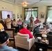 Military personnel in tan/khaki uniforms are conducting a presentation or visit at what appears to be a senior living facility or assisted living community. A service member stands near a display screen while addressing a group of elderly residents seated in a comfortable lounge-style room. The setting suggests a community outreach or veterans appreciation event.