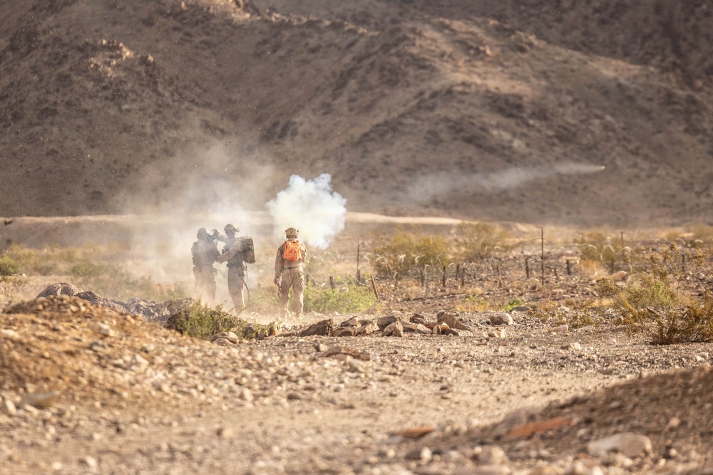 The image shows a small group of military personnel in a desert/arid terrain environment with rocky ground and barren hills in the background. One individual is wearing a distinctive orange vest or pack, suggesting a safety observer or controller role, while others appear in standard camouflage gear. A plume of white smoke is visible, consistent with a smoke grenade or pyrotechnic device deployment during what appears to be a training exercise.