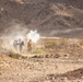 The image shows a small group of military personnel in a desert/arid terrain environment with rocky ground and barren hills in the background. One individual is wearing a distinctive orange vest or pack, suggesting a safety observer or controller role, while others appear in standard camouflage gear. A plume of white smoke is visible, consistent with a smoke grenade or pyrotechnic device deployment during what appears to be a training exercise.