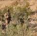 Two military personnel in desert camouflage uniforms are positioned in an arid, scrubland terrain. The foreground soldier is clearly visible wearing full combat gear including helmet, body armor, and carrying a weapon. A second soldier is visible in the background, slightly out of focus.
