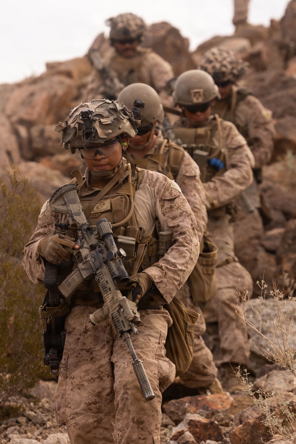 A file of military personnel in desert camouflage (MARPAT pattern) navigate rocky terrain in a arid environment. The lead soldier carries an M4/M16-style rifle with optics and is equipped with full combat gear including helmet, plate carrier, and tactical accessories. The image depicts what appears to be a patrol or training exercise in a desert rocky landscape.
