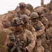 A file of military personnel in desert camouflage (MARPAT pattern) navigate rocky terrain in a arid environment. The lead soldier carries an M4/M16-style rifle with optics and is equipped with full combat gear including helmet, plate carrier, and tactical accessories. The image depicts what appears to be a patrol or training exercise in a desert rocky landscape.