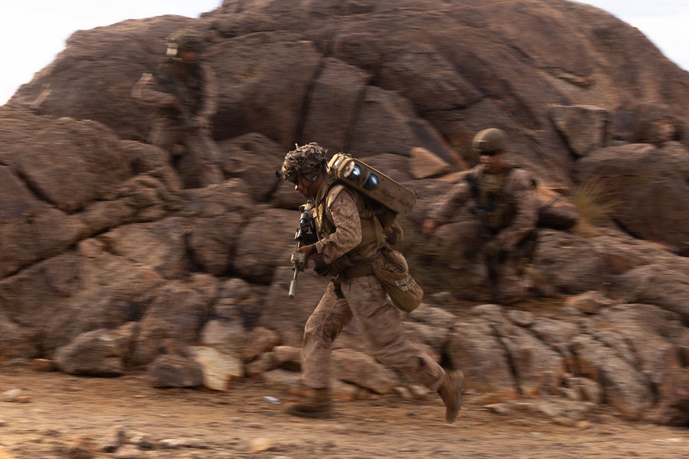 The image shows military personnel in desert camouflage uniforms moving rapidly across rocky, arid terrain. At least three soldiers are visible, with the foreground subject running while carrying a weapon and a large cylindrical object on their back. The motion blur effect suggests fast movement and conveys a sense of urgency consistent with a tactical field exercise or operation.