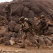 The image shows military personnel in desert camouflage uniforms moving rapidly across rocky, arid terrain. At least three soldiers are visible, with the foreground subject running while carrying a weapon and a large cylindrical object on their back. The motion blur effect suggests fast movement and conveys a sense of urgency consistent with a tactical field exercise or operation.