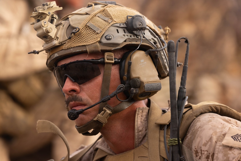 A close-up photograph of a male military personnel wearing full tactical gear in an arid, desert-like environment. The subject is equipped with advanced communications equipment, protective eyewear, and a tactical helmet with multiple attachments. A rank insignia is visible on the right shoulder of his uniform.