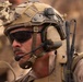 A close-up photograph of a male military personnel wearing full tactical gear in an arid, desert-like environment. The subject is equipped with advanced communications equipment, protective eyewear, and a tactical helmet with multiple attachments. A rank insignia is visible on the right shoulder of his uniform.