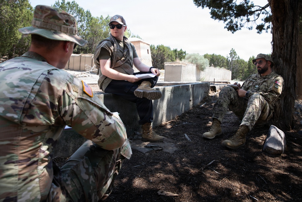 Three military personnel are gathered in an outdoor briefing or discussion near concrete barriers and trees. One individual wearing civilian-style tactical gear with a baseball cap and sunglasses appears to be leading the discussion while holding documents. The group includes at least one US Army soldier (identifiable by the American flag patch) and one soldier with a Canadian flag patch.