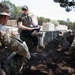 Three military personnel are gathered in an outdoor briefing or discussion near concrete barriers and trees. One individual wearing civilian-style tactical gear with a baseball cap and sunglasses appears to be leading the discussion while holding documents. The group includes at least one US Army soldier (identifiable by the American flag patch) and one soldier with a Canadian flag patch.