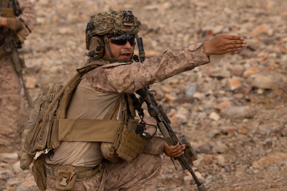 A heavily equipped soldier in desert camouflage (MARPAT pattern) is giving a hand signal or directing personnel while holding a rifle in a rocky, arid terrain. The soldier is wearing full combat gear including a tactical helmet with netting, communications headset, body armor, and a backpack. The environment and uniform pattern suggest a desert operational environment.