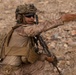 A heavily equipped soldier in desert camouflage (MARPAT pattern) is giving a hand signal or directing personnel while holding a rifle in a rocky, arid terrain. The soldier is wearing full combat gear including a tactical helmet with netting, communications headset, body armor, and a backpack. The environment and uniform pattern suggest a desert operational environment.