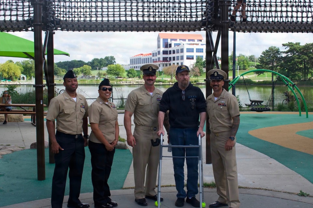 Five individuals pose for a group photo at an outdoor playground or park area near a body of water. Four of the individuals are wearing U.S. Navy uniforms in tan/khaki service dress, while one civilian man in the center uses a walker and wears a Navy-themed cap and shirt. A modern multi-story building is visible in the background across the water.