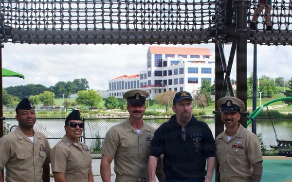 Five individuals pose for a group photo at an outdoor playground or park area near a body of water. Four of the individuals are wearing U.S. Navy uniforms in tan/khaki service dress, while one civilian man in the center uses a walker and wears a Navy-themed cap and shirt. A modern multi-story building is visible in the background across the water.
