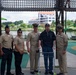 Five individuals pose for a group photo at an outdoor playground or park area near a body of water. Four of the individuals are wearing U.S. Navy uniforms in tan/khaki service dress, while one civilian man in the center uses a walker and wears a Navy-themed cap and shirt. A modern multi-story building is visible in the background across the water.