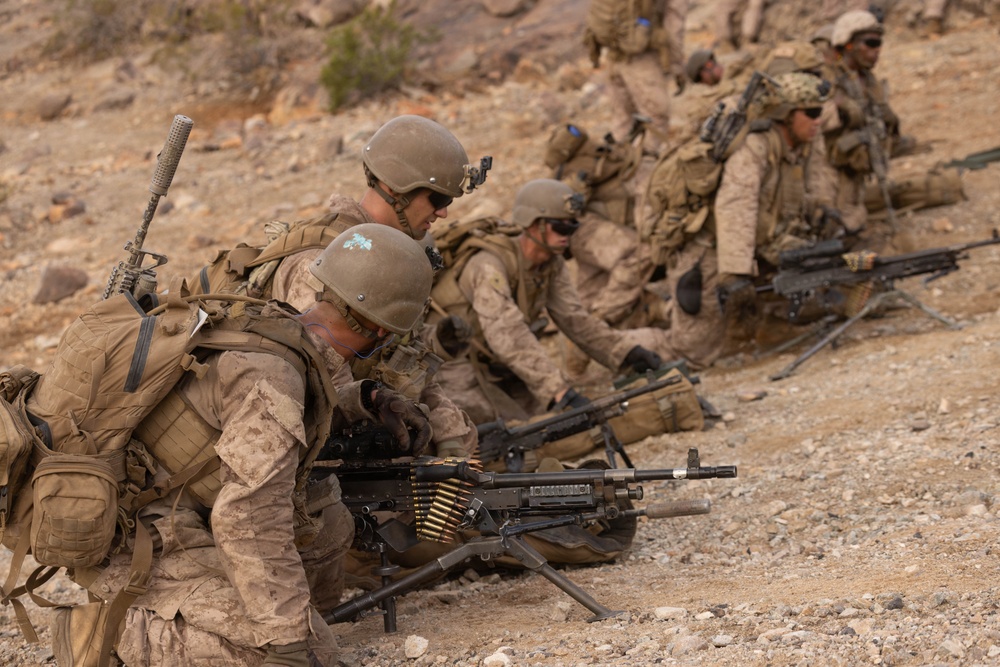 A line of military personnel in desert camouflage uniforms are positioned along a rocky, arid terrain in what appears to be a training exercise or tactical operation. The soldiers are equipped with heavy weapons, body armor, helmets, and tactical gear. The foreground prominently features a belt-fed machine gun mounted on a bipod with visible ammunition.