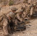 A line of military personnel in desert camouflage uniforms are positioned along a rocky, arid terrain in what appears to be a training exercise or tactical operation. The soldiers are equipped with heavy weapons, body armor, helmets, and tactical gear. The foreground prominently features a belt-fed machine gun mounted on a bipod with visible ammunition.