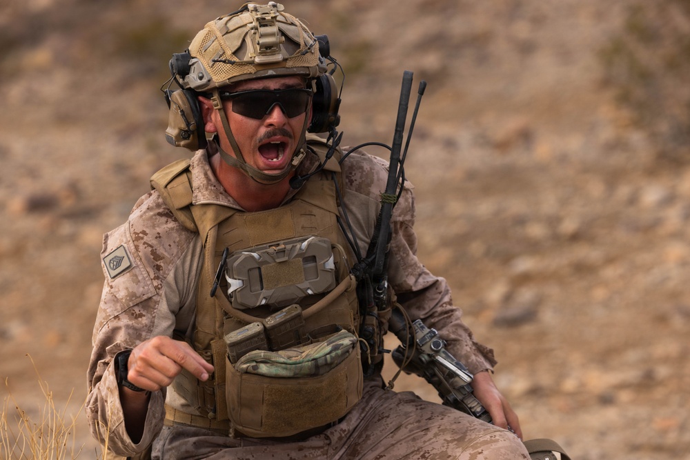 A U.S. Marine in full combat gear is captured mid-shout in an arid, desert-like training or operational environment. He is wearing MARPAT desert camouflage, a tactical vest, ballistic helmet with communications equipment, and protective eyewear. His expression and posture suggest he is issuing commands or responding urgently to a situation.