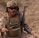 A U.S. Marine in full combat gear is captured mid-shout in an arid, desert-like training or operational environment. He is wearing MARPAT desert camouflage, a tactical vest, ballistic helmet with communications equipment, and protective eyewear. His expression and posture suggest he is issuing commands or responding urgently to a situation.