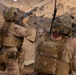 Two military personnel in desert camouflage uniforms are engaged in a tactical exercise in an arid, rocky terrain. The soldier on the left is aiming a shotgun upward while the soldier on the right, identified by name tape 'ADLEY' and helmet marking 'G4B', appears to be communicating or calling out. Both are equipped with full tactical gear including body armor, helmets, and radio antennas.