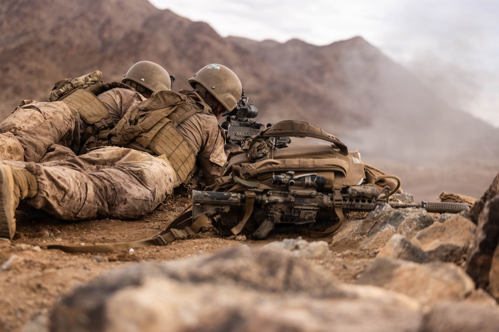 Two soldiers in desert camouflage uniforms are in a prone firing position on rocky, arid terrain. They are equipped with tactical gear, helmets, body armor, and rifles, with one soldier actively aiming through a scope. The background shows a mountainous desert landscape with dust or smoke visible in the distance.