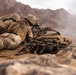 Two soldiers in desert camouflage uniforms are in a prone firing position on rocky, arid terrain. They are equipped with tactical gear, helmets, body armor, and rifles, with one soldier actively aiming through a scope. The background shows a mountainous desert landscape with dust or smoke visible in the distance.